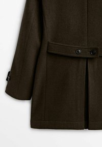 Cappotto di lana marrone scuro con una vestibilità strutturata, dotato di una vita con cintura, chiusura a doppio bottone e spacco laterale per facilitare i movimenti.