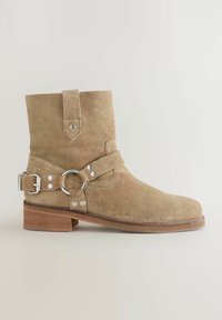 Stivaletto in suede beige chiaro con punta arrotondata, cinturino regolabile con anello metallico e tacco a blocco in legno.
