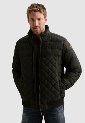 Homme aux cheveux courts portant une veste matelassée vert foncé, les mains dans les poches, debout devant un fond uni.