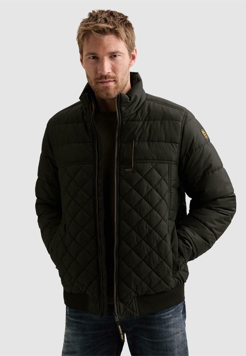 Homme aux cheveux courts portant une veste matelassée vert foncé, les mains dans les poches, debout devant un fond uni.