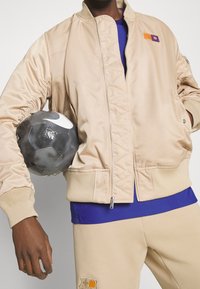 Beige Bomberjacke mit gerippten Bündchen und gestepptem Vorderteil, kombiniert mit einem blauen Hemd und beigen Hosen mit Logo, während er einen schwarzen Fußball hält.