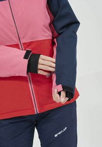 Giacca da sci colorata con pannelli rosa, rossi e blu navy; chiusure a zip e Velcro; il tessuto appare liscio con una finitura opaca; polsino regolabile a mano.