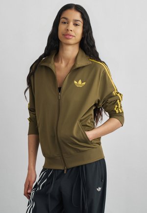 Junge Frau mit langen dunklen Haaren, die eine olivgrüne Jacke mit Reißverschluss und gelben Streifen sowie schwarze Adidas-Trainingshose mit weißen Streifen trägt.