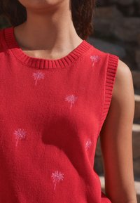 Detailný záber osoby oblečenej v červenom pletenom topánke bez rukávov s malými bielymi vzormi palmových stromov, vonku na slnku.