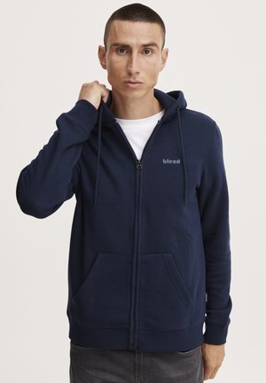 Mornarsko modra zip-up kapuca iz mehkega materiala, z sprednjim žepom, kapuco z vrvico in logotipom na prsih. Gladka tekstura, sproščen kroj.