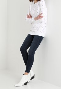 Persona che indossa un sudore bianco a motivi, jeans skinny scuri e stivaletti bianchi lucidi con tacco angolato, appoggiata a un muro bianco.