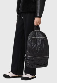 Zaino nero trapuntato con zip argentate, caratterizzato da una superficie testurizzata e da una maniglia superiore, tenuto da una persona vestita di nero.
