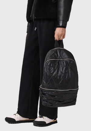 Zaino nero trapuntato con zip argentate, caratterizzato da una superficie testurizzata e da una maniglia superiore, tenuto da una persona vestita di nero.