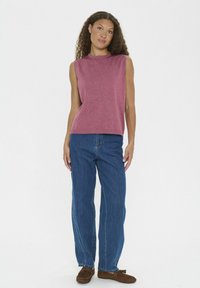 Frau mit lockigem Haar steht in einem ärmellosen rosa Oberteil, blue Jeans und braunen Mokassins vor einem weißen Hintergrund.