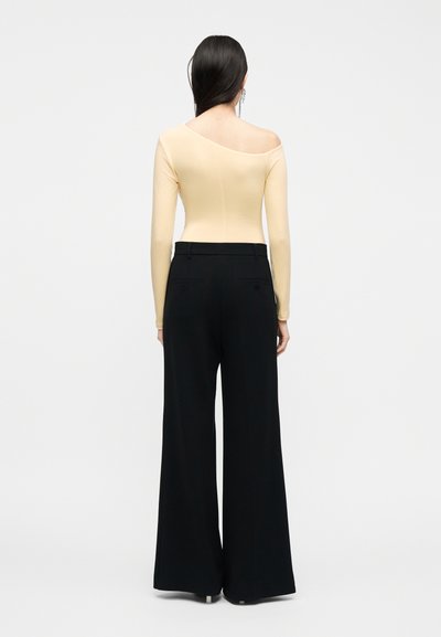 Γυναίκα με μακριά μαύρα μαλλιά φοράει μπεζ τοπ με μακριά μανίκια και στενό λαιμόκοψη, μαζί με μαύρο παντελόνι φαρδιάς ποδός, στέκεται μπροστά σε απλό φωτεινό φόντο.