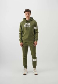 Olivgrünes Hoodie- und Jogginganzug-Set mit weißem Logo und Streifenakzenten, ausgestattet mit einer Kapuze mit Kordelzug und gerippten Bündchen, kombiniert mit weißen Sneakers.