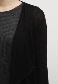 Cárdigan negro ligero con un diseño abierto y drapeado; combinado con una parte superior gris texturizada que presenta costillas diagonales.