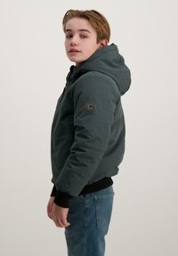 Groene hoodie met een gladde textuur, ritssluiting, geribbelde manchetten en een logo-opdruk op de linkermouw. Draagt over een zwarte top en jeans.