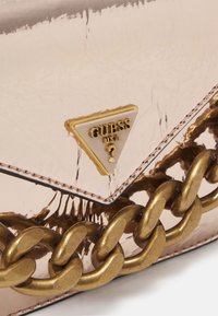 Angolo di una borsa lucida color oro rosa con tracolla in catena dorata e logo triangolare Guess USA con punto interrogativo.