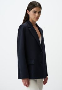 Blazer blu navy realizzato in tessuto testurizzato, con un design a un petto, rever a punta e taschino frontale. I bottoni sono neri.