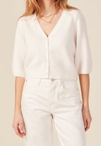 Cardigan bianco corto con maniche a metà, chiusura a bottoni, bordo a coste, realizzato in materiale morbido. Indossato sopra jeans bianchi a vita alta.