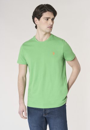 Giovane uomo con capelli castani che indossa una maglietta a maniche corte di un verde brillante e jeans blu, che guarda a sinistra con le mani rilassate.