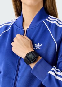 Juodas skaitmeninis laikrodis su mėlynu ekranu ant riešo, derinamas su mėlynu sportiniu švarku, turinčiu baltas juosteles ir Adidas logotipą.