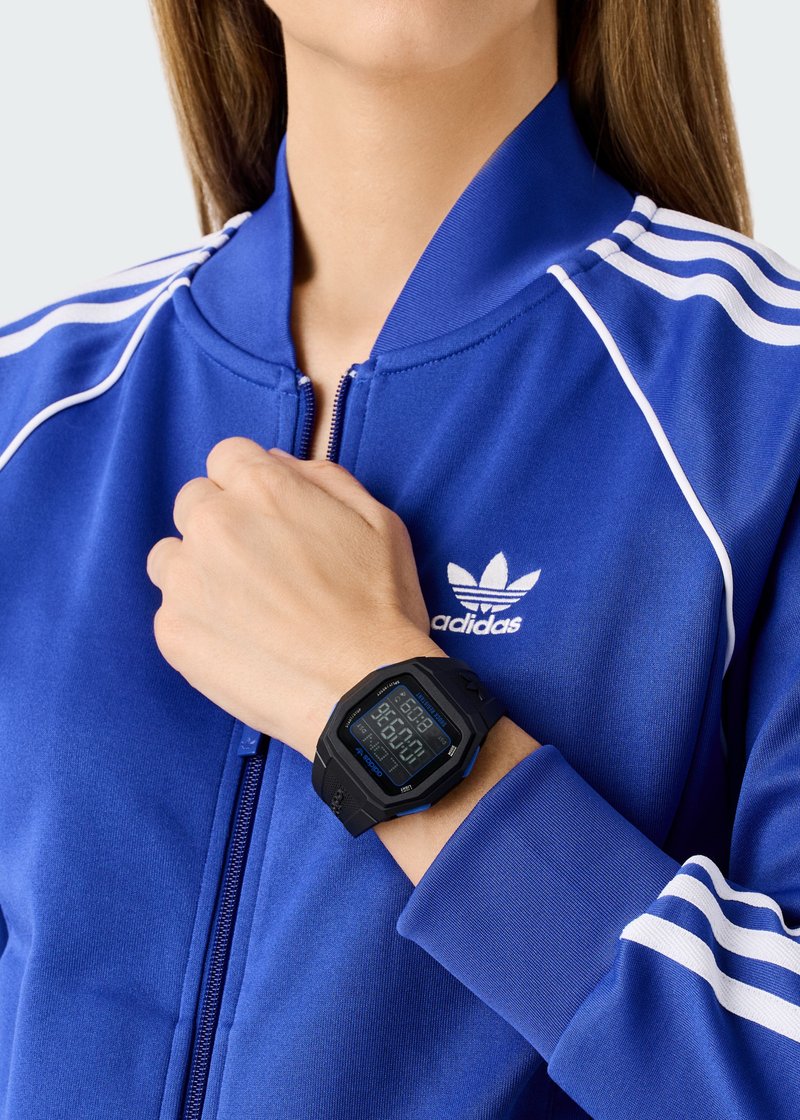 Juodas skaitmeninis laikrodis su mėlynu ekranu ant riešo, derinamas su mėlynu sportiniu švarku, turinčiu baltas juosteles ir Adidas logotipą.