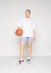 Baltas per didelis marškinėliai su mėlynu logotipu, šviesiai violetinės šortai ir juodi/rožiniai sportbačiai, laikantys krepšinį. Slygi medžiaga ir laisvas sijonas.