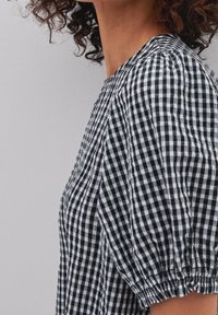 Kvinna i blus med puffärmar i svart och vitt rutigt mönster, med samlade detaljer vid ärmsluten och halsringningen, synligt lockigt hår.