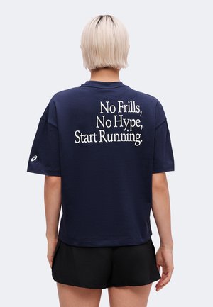 Person med kort blondt hår iført en navy trøje med teksten "No Frills, No Hype, Start Running" og sorte shorts set bagfra.