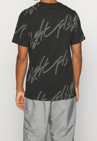 Homme portant un t-shirt noir à manches courtes avec des motifs de texte graphique et de logo gris, associé à un pantalon léger gris ample, vu de dos.