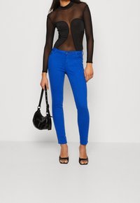Jeans ajustados azules con una textura suave, que cuentan con un cierre de botón en la parte delantera y cinco bolsillos, combinados con una blusa negra de manga larga y transparente.