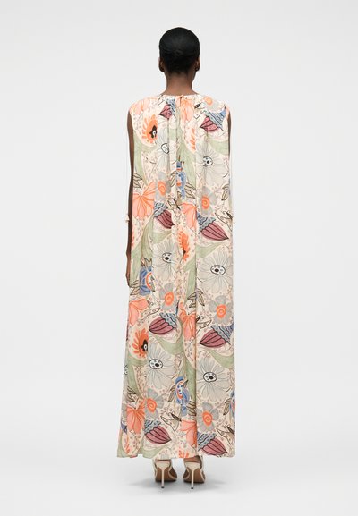 Γυναίκα που στέκεται φορώντας ένα αμάνικο, μακρύ φόρεμα μέχρι το πάτωμα με πολύχρωμο λουλουδάτο σχέδιο, σε συνδυασμό με μπεζ ψηλοτάκουνα σανδάλια.