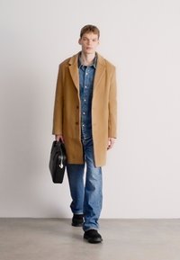 Καφέ μάλλινο παλτό, τζιν πουκάμισο, μπλε τζιν και μαύρα παπούτσια. Το μοντέλο κρατά μια μαύρη χαρτοφάκο, στέκεται σε ένα γκρι δάπεδο μπροστά σε έναν ανοιχτό φόντο.