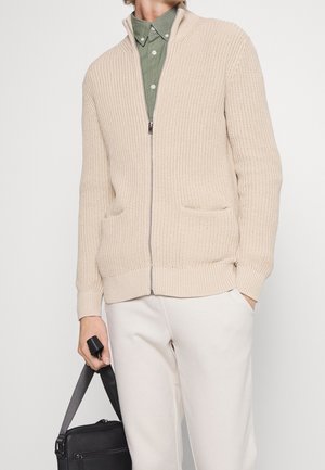 Beige gebreide zip-up cardigan met ribbelstructuur, zijzakken en een hoge kraag. Gedragen over een groene button-up shirt.