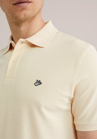 Gaiši beidzēts kokvilnas polo krekls ar teksturētu rakstu, ar trim pogām un melnu izbrodētu putna logo kreisajā krūtī.