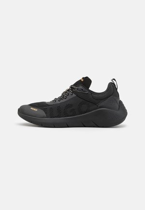 Baskets de sport noires avec empeigne en mesh, lacets à motifs et semelle épaisse, marquées de "Hugo" en lettres dorées sur le côté et la languette.