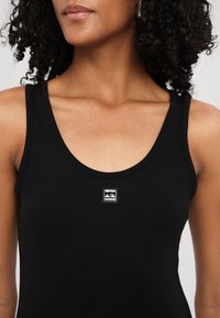 Femme portant un débardeur noir avec un petit logo carré représentant deux voiliers blancs sur la poitrine.