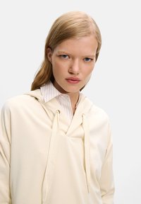 Cream hoodie with a front pocket, soft fabric, and a drawstring hood over a light striped collared shirt. Smooth texture and relaxed fit.