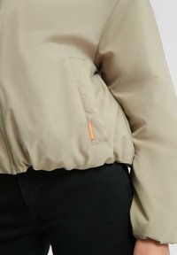 Doudoune beige avec poche latérale et étiquette orange, portée avec un pantalon noir, gros plan sur la taille et le bras.