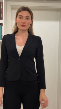 Γυναίκα με ίσια ανοιχτόκαστα μαλλιά που φοράει μαύρο σακάκι πάνω από λευκή μπλούζα, στέκεται σε εσωτερικό χώρο μπροστά σε λευκό τοίχο και φόντο ραφιέρας βιβλίων.