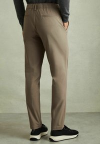Pantaloni bej, croiți cu un aspect neted, având două buzunare la spate și un croi conic, asociați cu teniși sport negri.