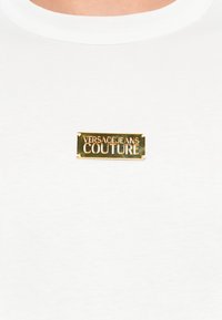 Λευκό βαμβακερό μπλουζάκι με ορθογώνια χρυσή μεταλλική ετικέτα, εντυπωμένη με τη γραφή "VERSACEJEANS COUTURE" σε έντονη γραμματοσειρά, τοποθετημένη στο στήθος.