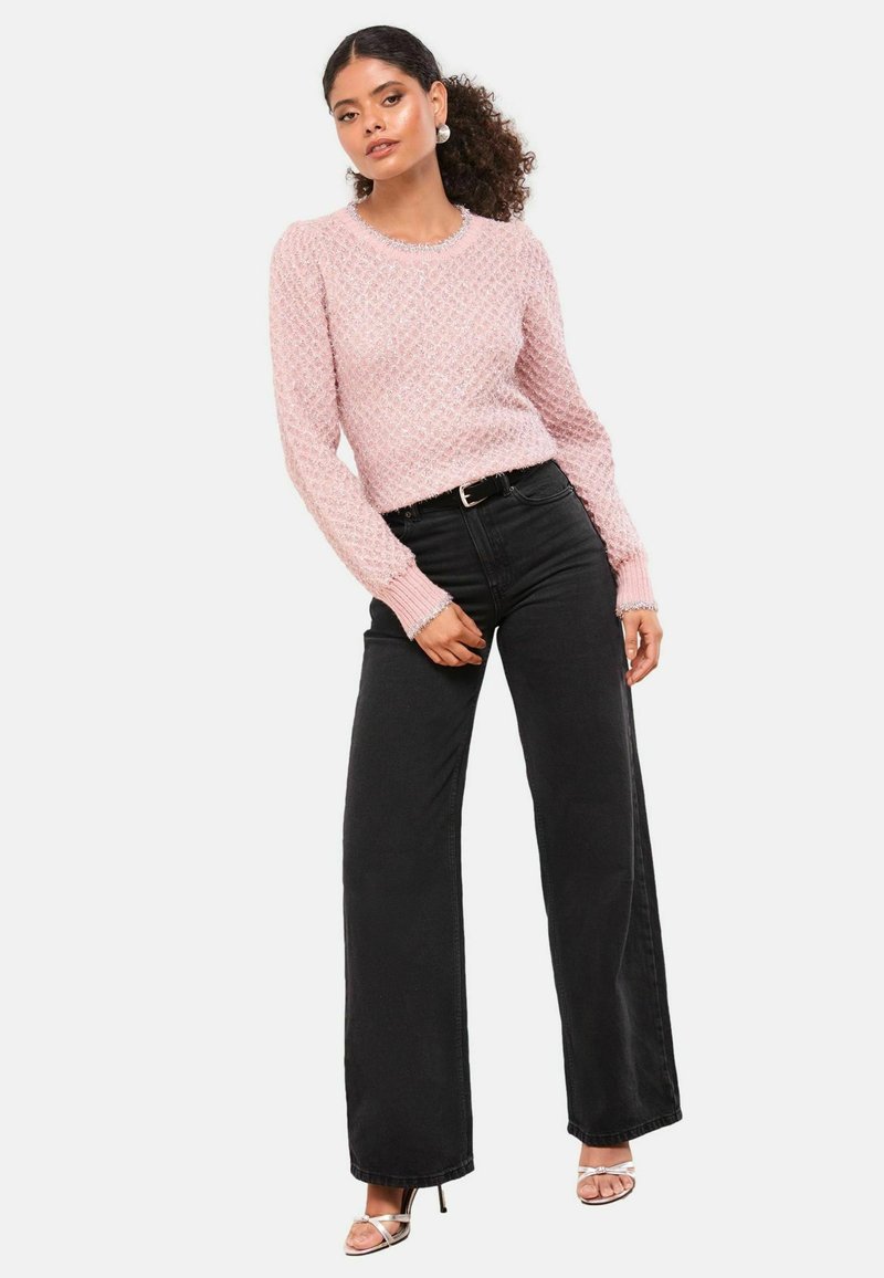 Rózsaszín kötött pulóver, texturált gyémántmintával, fekete, magas derekú, széles szárú farmerral párosítva. A modellen ezüst színű magassarkú szandál található.