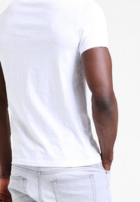 Bílá, krátká tričko s hladkou texturou a přiléhavým střihem, v kombinaci se světle šedými kalhotami s zadními kapsami a dvojitým švem.