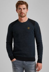 Granatowy sweter o teksturowanej powierzchni z raglanowymi rękawami, ściągaczami przy mankietach i u dołu, z małym żółtym logo na klatce piersiowej oraz kontrastującymi czarnymi akcentami na ramionach.