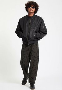 Must jopetükk esiosal tõmblukuga, rinnal tikanddetail, seljas tumerohelised püksid nahkhiire mustriga ja mustad kingad.