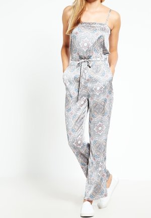 Kobieta w bezrękawnikowym kombinezonie w wzory, z elastycznym pasem, z rękami w kieszeniach i białych butach slip-on, stojąca na jasnym tle.