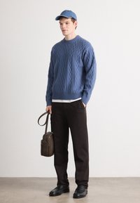 Blauwe kabelbrei trui, zwarte broek en zwarte schoenen. Accessoires zijn een blauwe pet en een bruine leren schoudertas.