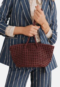 Vävd bourgognehandväska med dubbla handtag, med en texturerad, öppnarbetad design. Står mot en randig blå kostym med vita accenter.