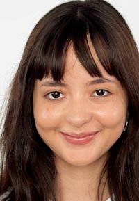 Photo en gros plan d'une personne avec de longs cheveux brun foncé et une frange, portant une boucle d'oreille en argent. Teint lisse, arrière-plan neutre.