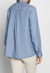 Light blue denim shirt with long sleeves, button cuffs, and a pleated back design. Rounded hem and smooth texture.