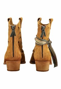 Dos botines de ante color tostado con cremalleras traseras, tacones de madera y un botín atado con una cinta de tela a rayas.