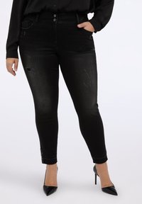 Jeans negros ajustados con un ligero desgaste, cierre de dos botones y una textura elegante. Presenta herrajes decorativos en el tobillo.